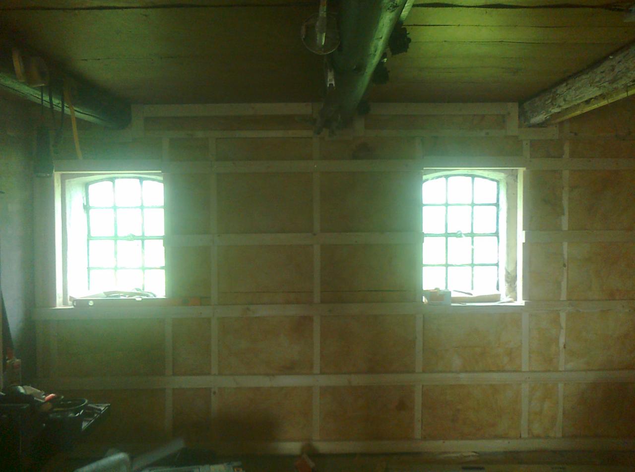 Wnętrze remontowanego pomieszczenia z dwoma oknami, ściany wyłożone płytami i drewnianą konstrukcją. Widoczne narzędzia na pierwszym planie.