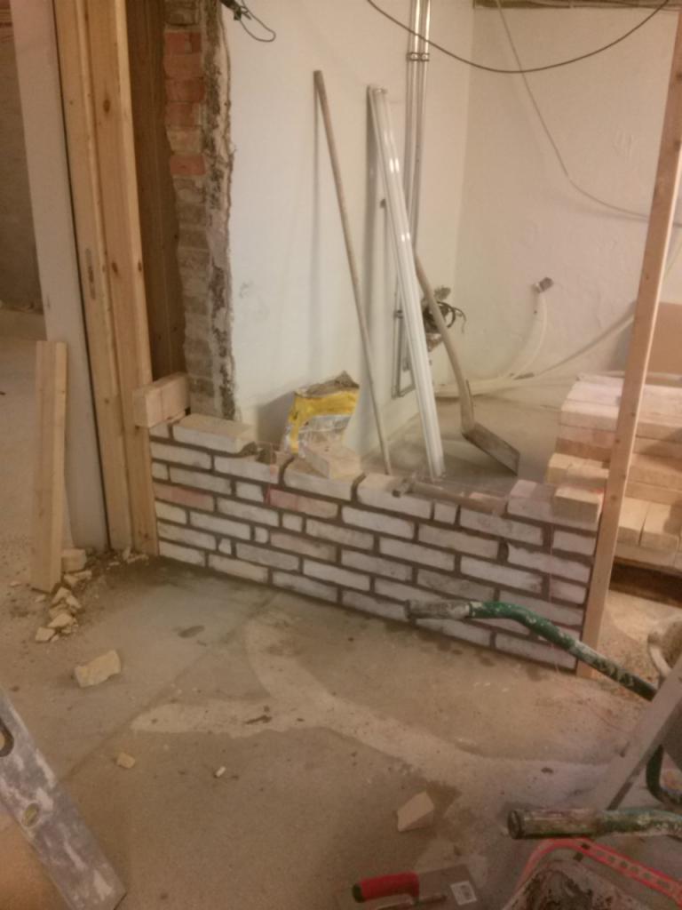 Murowanie ściany z białej cegły w trakcie remontu. Widoczne narzędzia, zaprawa i fragmenty starej ściany. Surowy stan, oświetlenie robocze.