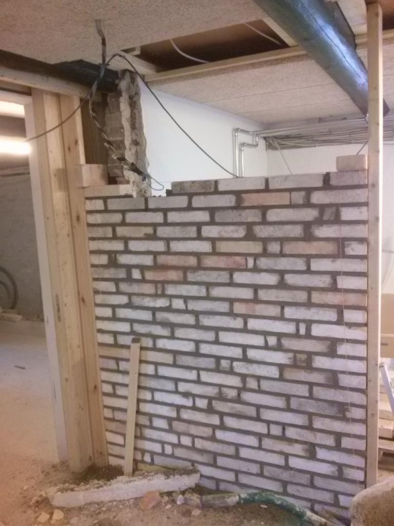 Wznoszenie muru z cegły wewnątrz budynku. Widoczne przewody instalacyjne, drewniane elementy konstrukcyjne i surowy stan wykończenia wnętrza.
