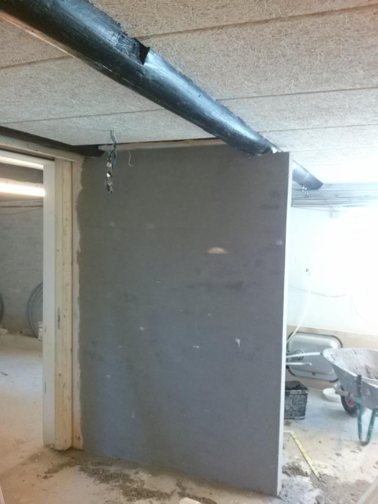 Szara ściana w trakcie budowy/remontu, widoczne elementy konstrukcyjne i narzędzia, sufit z drewnianą belką. Ujęcie z bliska, wnętrze w stanie surowym.