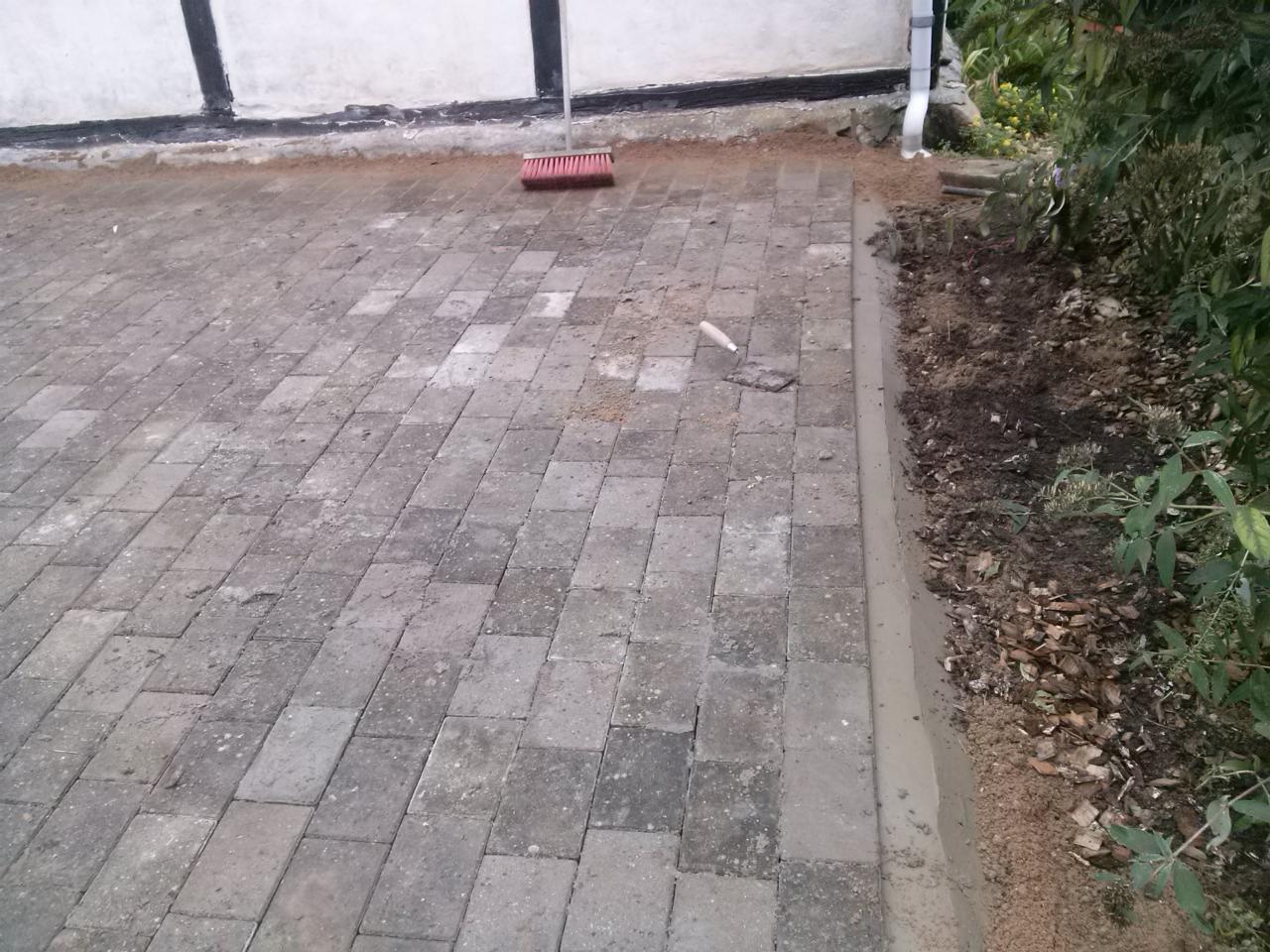 Ułożona kostka brukowa na posesji, widoczna szczotka i kielnia, świeżo obsypane piaskiem, obrzeża z betonu, roślinność wzdłuż krawędzi.