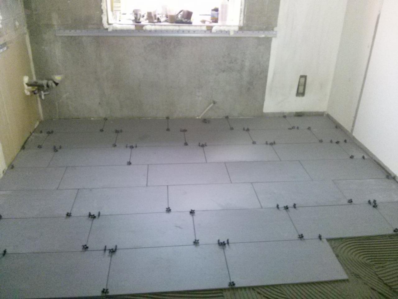 Szare płytki podłogowe w trakcie układania, widoczne krzyżyki dystansowe i warstwa kleju z wyraźnym wzorem. Ściany przygotowane do dalszych prac wykończeniowych.