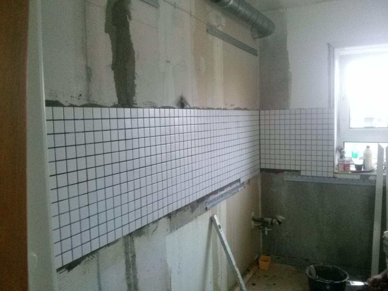 Wnętrze w trakcie wykańczania z częściowo ułożonymi białymi, kwadratowymi płytkami na ścianach. Widoczne narzędzia i surowy stan ścian.