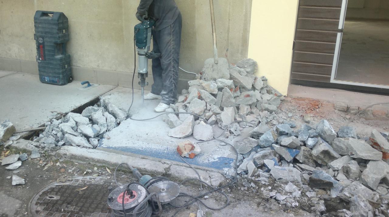 Pracownik w trakcie wyburzania betonowej posadzki przy budynku za pomocą młota pneumatycznego. Obok leżą gruz i narzędzia.