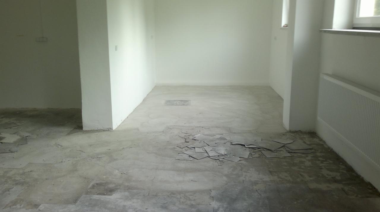 Wnętrze pokoju z resztkami starej podłogi i przygotowaną powierzchnią pod nową. Widoczne fragmenty płytek i białe ściany, grzejnik. Przygotowanie do remontu.