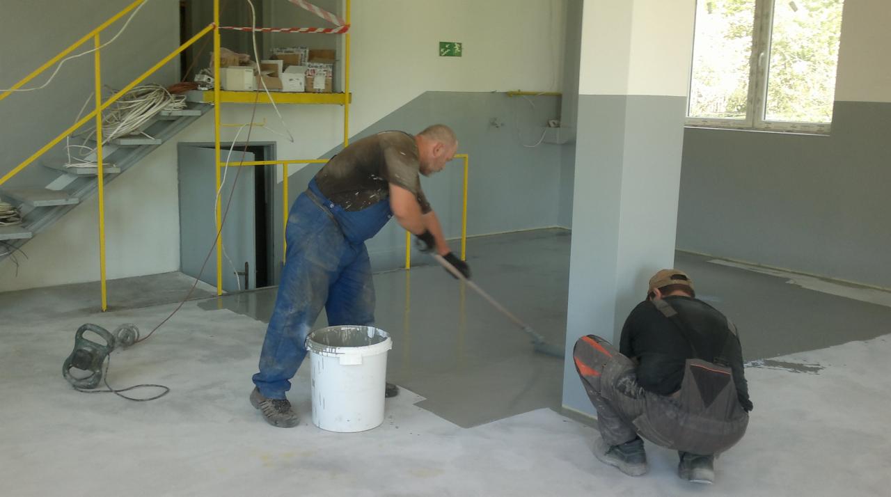 Malowanie podłogi w pomieszczeniu przemysłowym. Dwóch pracowników rozprowadza szarą farbę za pomocą wałka i pędzla. Widoczne schody, okno i narzędzia.