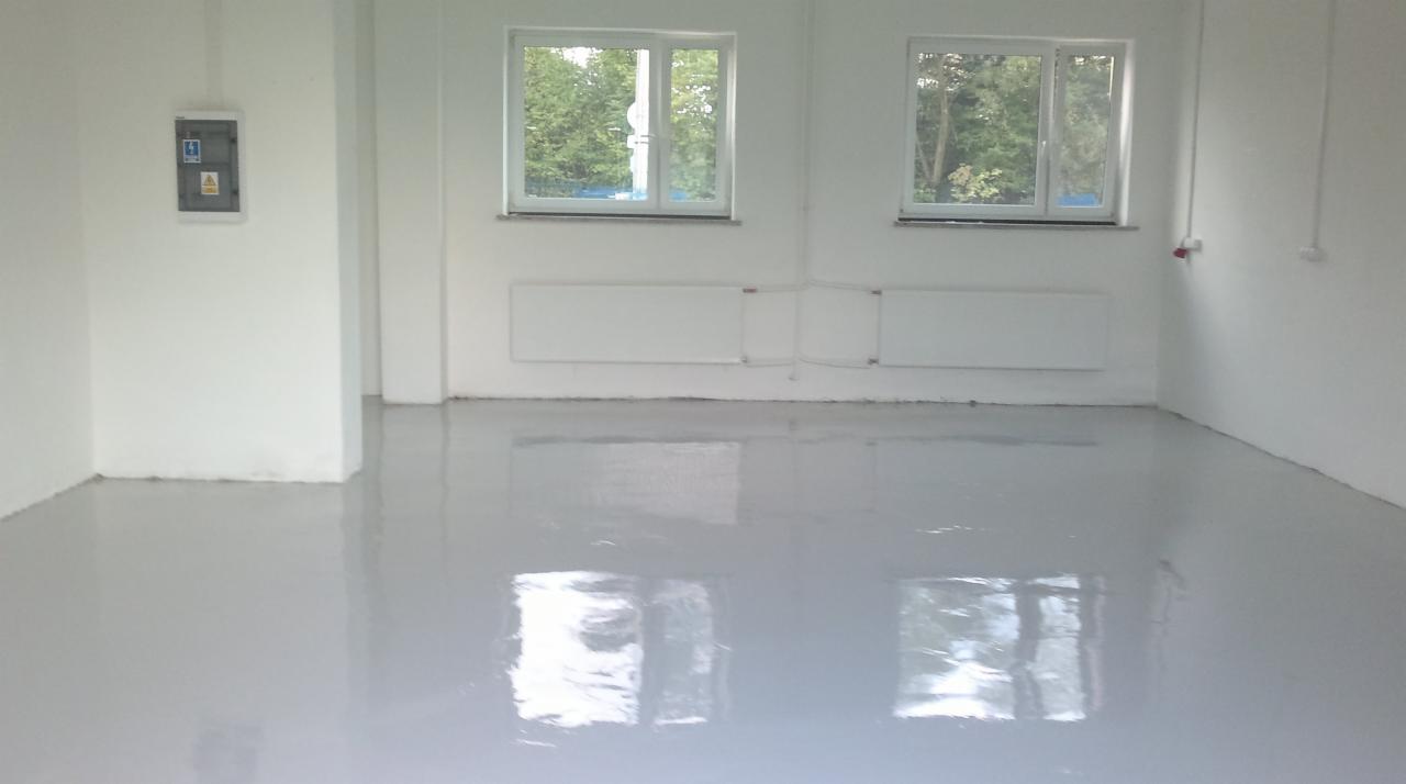 Wnętrze z szarą, błyszczącą posadzką żywiczną, białymi ścianami, grzejnikami i dwoma oknami z widokiem na zieleń. Na ścianie tablica elektryczna.