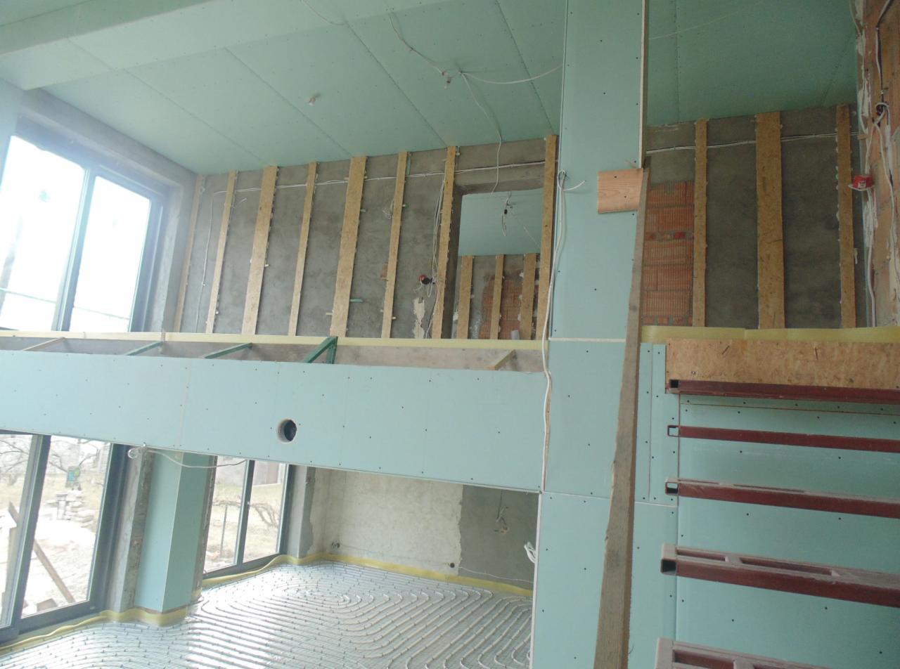 Wnętrze w trakcie prac wykończeniowych: płyty gipsowe na ścianach i suficie, instalacja ogrzewania podłogowego, drewniana konstrukcja. Widoczne okna i surowy stan ścian.