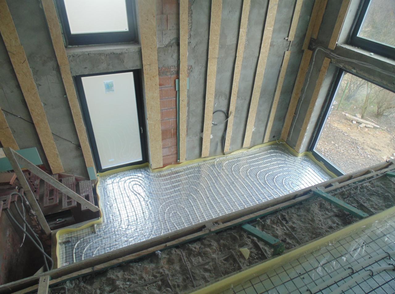 Instalacja ogrzewania podłogowego w surowym wnętrzu z oknami i przygotowanymi ścianami. Widoczne rury grzewcze ułożone na siatce, gotowe do zalania wylewką.