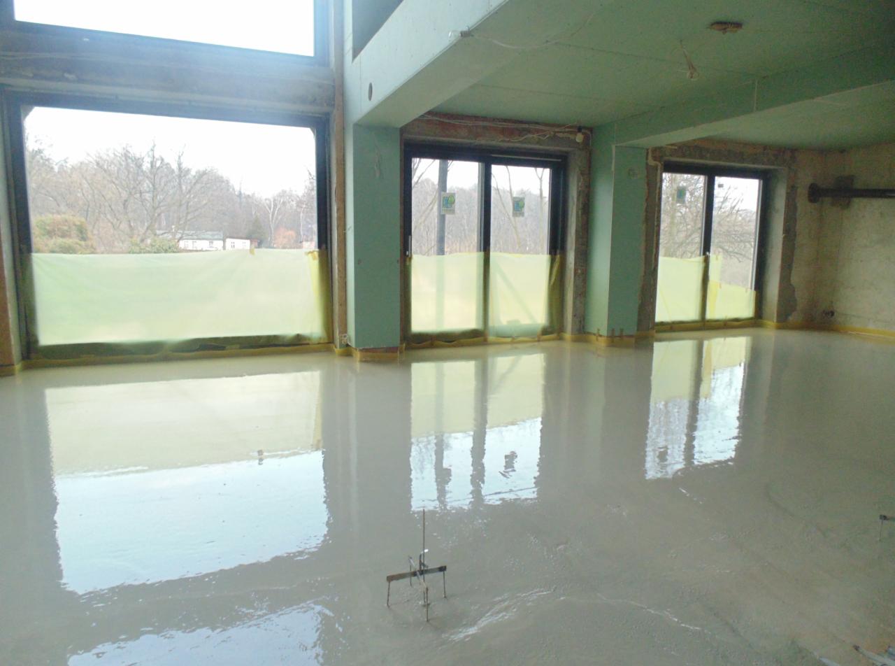 Wylany, mokry podkład podłogowy w jasnym pomieszczeniu z dużymi oknami zabezpieczonymi folią. Widoczne narzędzie do poziomowania w podkładzie.