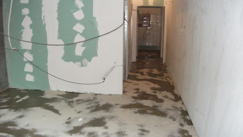 Pokój w trakcie szpachlowania: ściany z widocznymi łatami gipsowymi, mokra podłoga. Widoczne przewody i metalowe profile. W tle otwarte drzwi do kolejnego pomieszczenia.