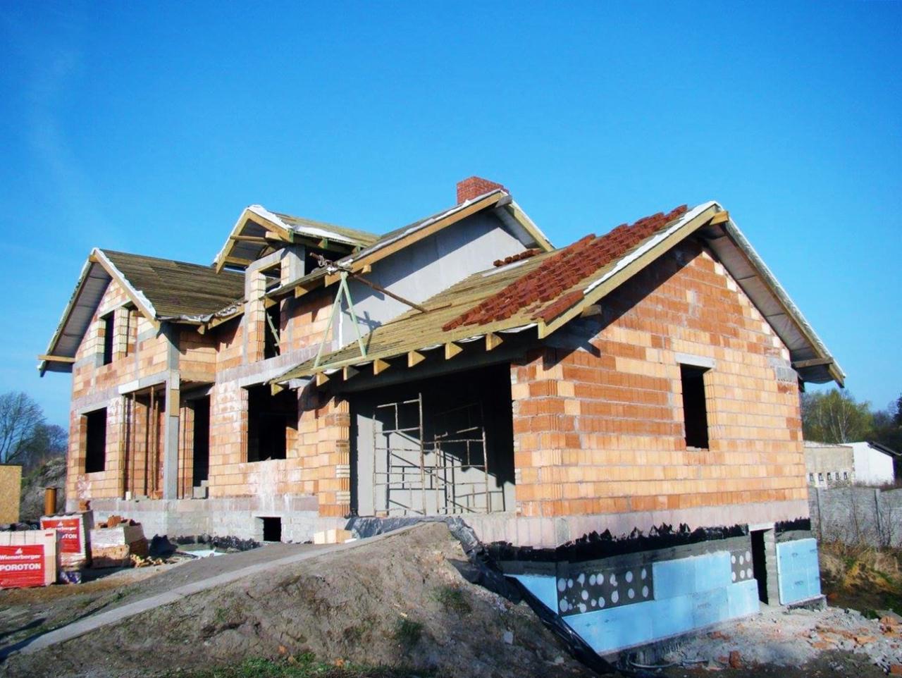 Niedokończony budynek jednorodzinny z czerwonej cegły Porotherm, częściowo zadaszony, z widocznym rusztowaniem i materiałami budowlanymi na placu budowy, na tle błękitnego nieba.