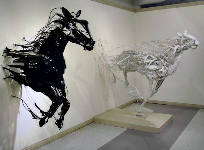 Dwie abstrakcyjne rzeźby koni: czarny koń przymocowany do ściany i biały stojący na podeście, wykonane z metalowych elementów w galerii.