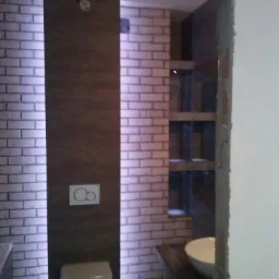 Wykończona łazienka z ceglastymi płytkami, podświetleniem LED, drewnianym panelem i półkami w ścianie. Toaleta zabezpieczona folią ochronną.