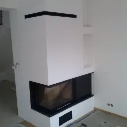 Wykończony kominek narożny z czarnym obramowaniem w jasnym wnętrzu w trakcie remontu w Zakliczynie, widoczne narzędzia na podłodze.