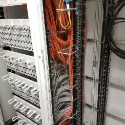 Rozdzielnica telekomunikacyjna z uporządkowanymi kablami światłowodowymi w kolorach pomarańczowym, żółtym, szarym i czarnym, zamontowanymi w metalowych korytkach i uchwytach na białej ścianie.