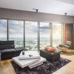 MANUFABRYKA STUDIO PROJEKTOWE KATARZYNA BURZYŃSKA - Nowoczesny salon z panoramicznym widokiem na miasto, szara sofa narożna z pomarańczowymi poduszkami, designerski stolik kawowy i fotel Eames Lounge Chair.