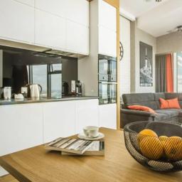 MANUFABRYKA STUDIO PROJEKTOWE KATARZYNA BURZYŃSKA - Nowoczesne wnętrze mieszkania z otwartą kuchnią, białymi szafkami, szarą sofą narożną z pomarańczowymi poduszkami i widokiem na miasto z okna.