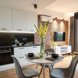 Jasna kawalerka z aneksem kuchennym: białe szafki, czarny blat, ceglana ściana z telewizorem, okrągły stół z szarymi krzesłami i wazon z żółtymi kwiatami.