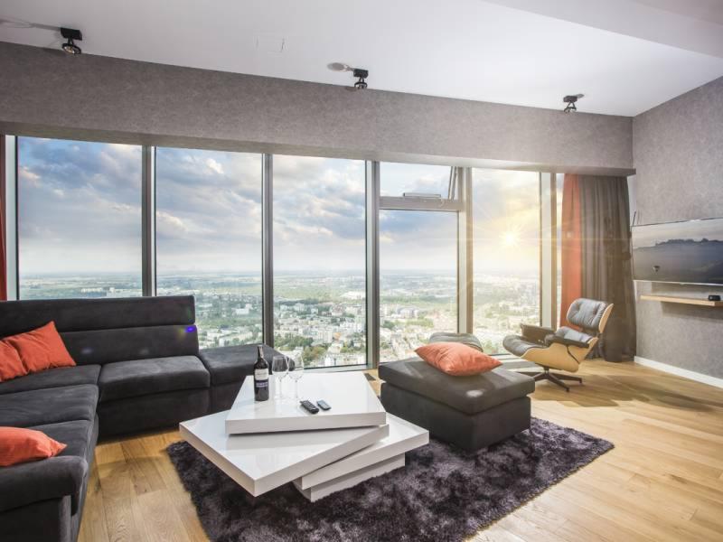 Nowoczesny salon z panoramicznym widokiem na miasto, szara sofa narożna z pomarańczowymi poduszkami, designerski stolik kawowy i fotel Eames Lounge Chair.