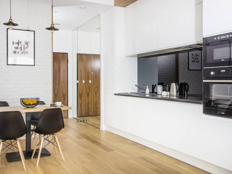 Jasne wnętrze z drewnianą podłogą, białymi szafkami kuchennymi, czarnym blatem, stołem z czarnymi krzesłami i plakatem motywacyjnym na ścianie z białej cegły.