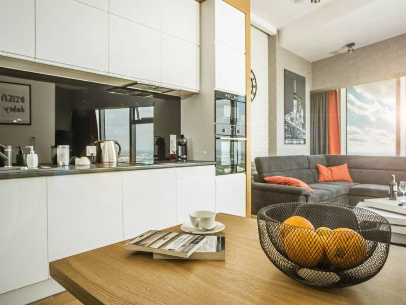 Nowoczesne wnętrze mieszkania z otwartą kuchnią, białymi szafkami, szarą sofą narożną z pomarańczowymi poduszkami i widokiem na miasto z okna.