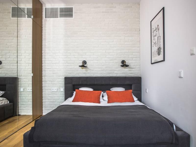 Sypialnia z łóżkiem z szarą tapicerką, pomarańczowymi poduszkami na białej pościeli, białą ścianą z cegły i czarnymi kinkietami, obok lustrzana szafa