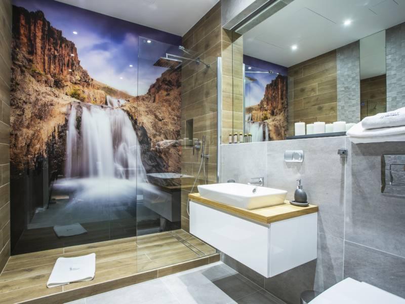 Nowoczesna łazienka z szklaną kabiną prysznicową i fototapetą z motywem wodospadu, biała umywalka nablatowa na drewnianym blacie, szare płytki, minimalistyczne akcesoria.