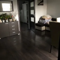 Uporządkowane wnętrze pokoju z ciemną podłogą, komodą z dekoracjami roślinnymi, otwartymi drzwiami do kolejnego pomieszczenia i stertą pościeli na kanapie w tle.