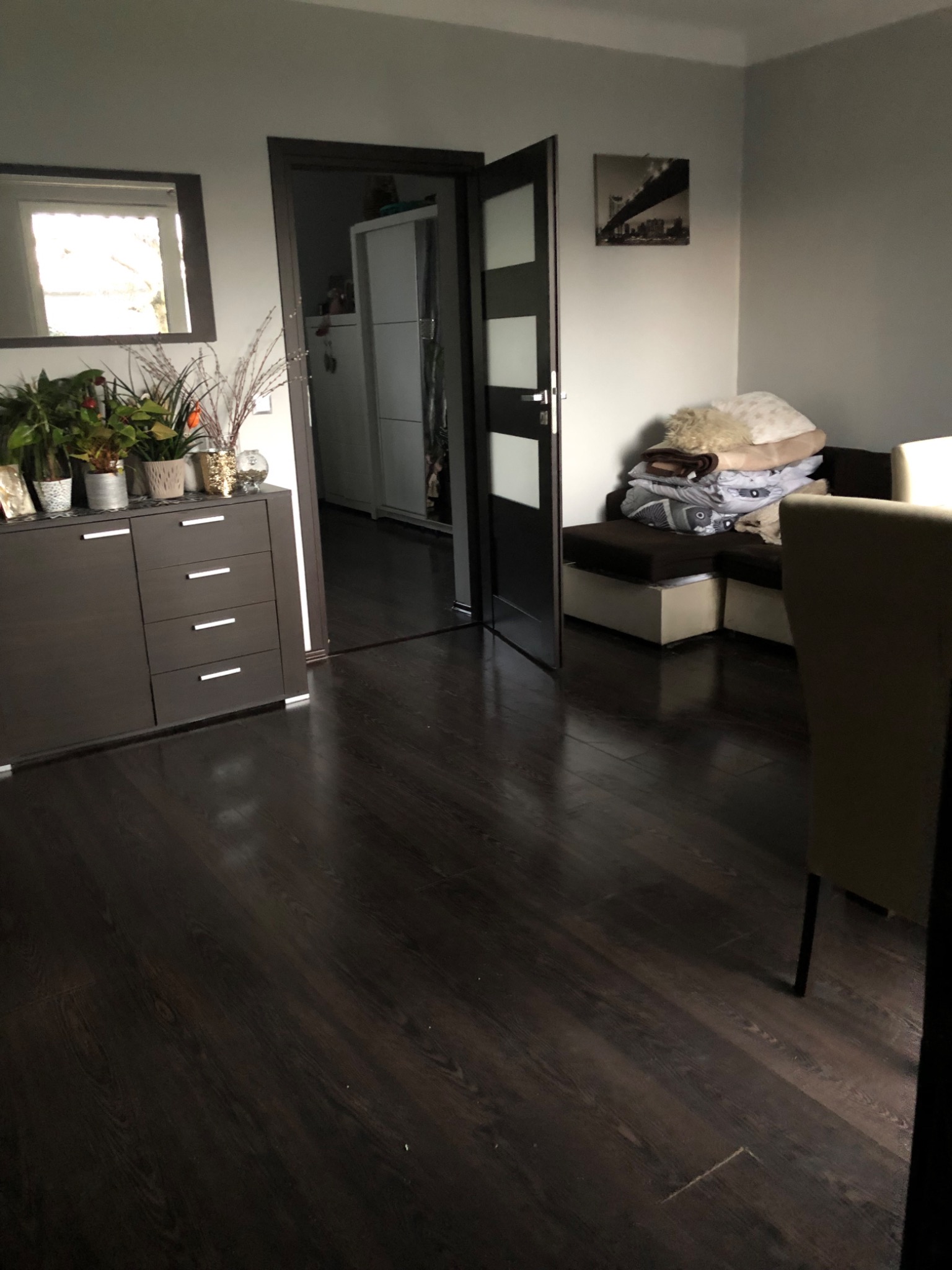 Uporządkowane wnętrze pokoju z ciemną podłogą, komodą z dekoracjami roślinnymi, otwartymi drzwiami do kolejnego pomieszczenia i stertą pościeli na kanapie w tle.