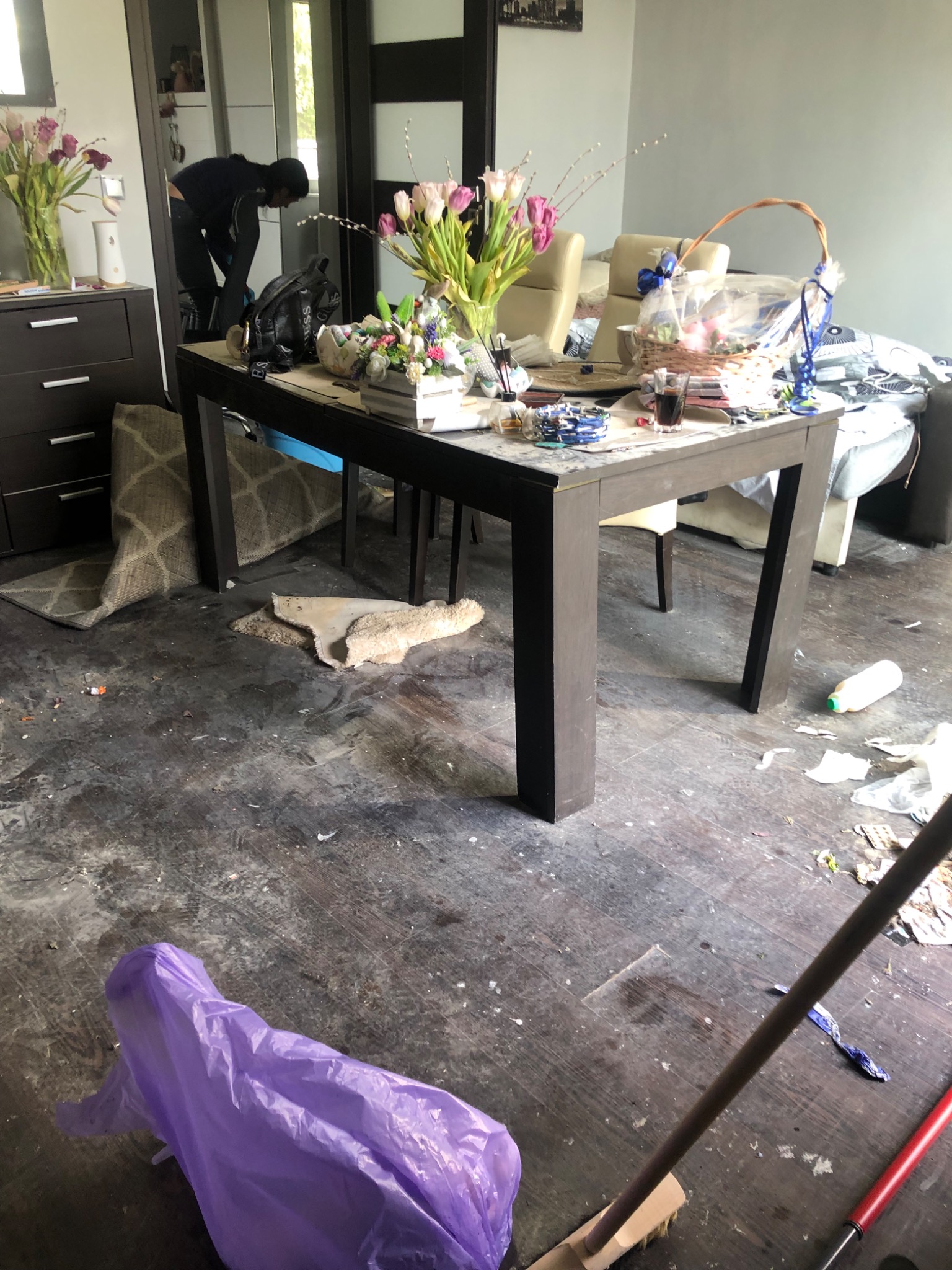 Widok na zabałaganione pomieszczenie z brudną podłogą, gdzie osoba w ciemnych ubraniach sprząta przy stole, a na podłodze leżą śmieci, worek foliowy i mop.
