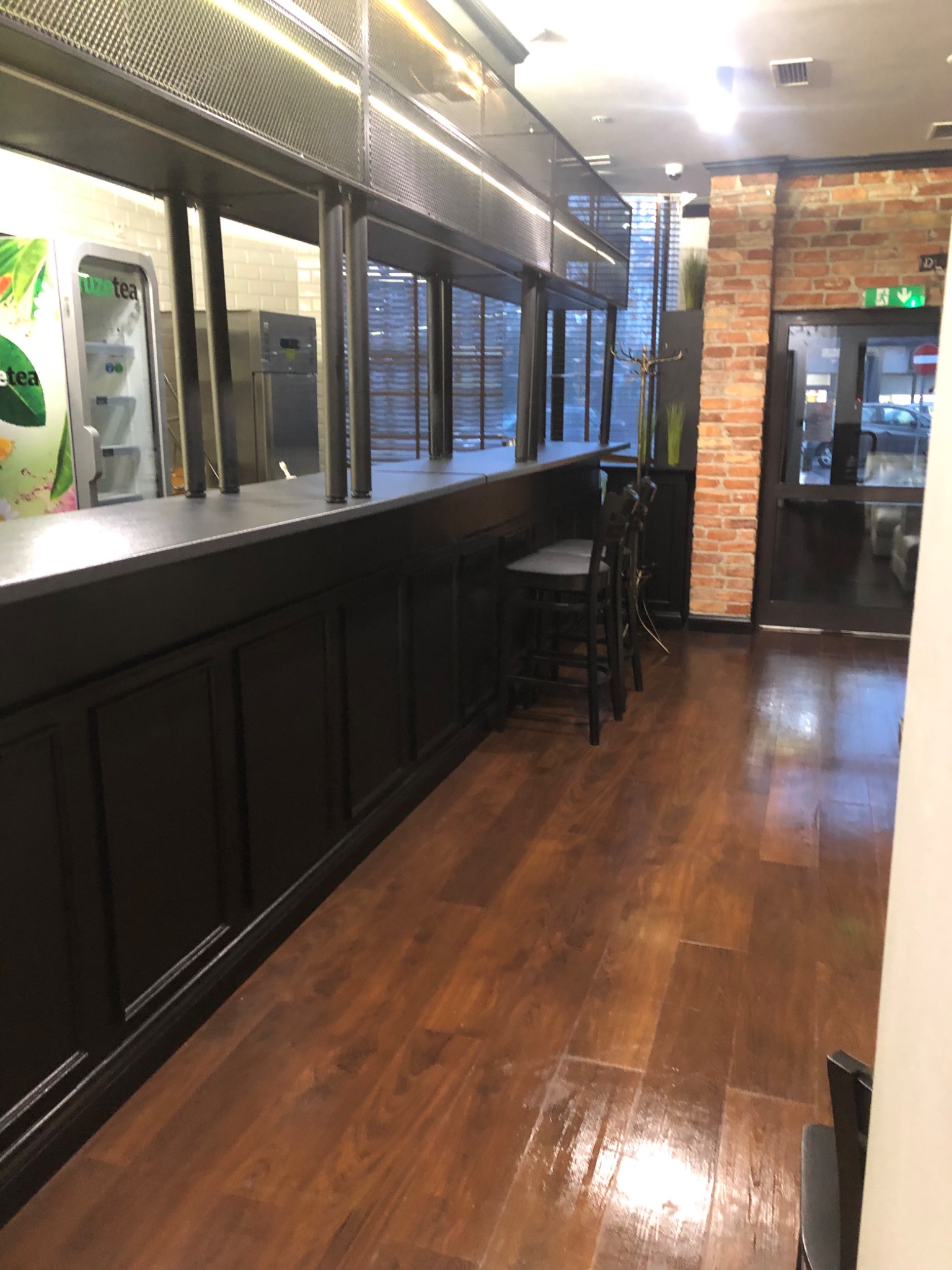 Wnętrze lokalu gastronomicznego z czarnym barem, metalową konstrukcją nad nim, drewnianą podłogą, ceglaną ścianą i widoczną lodówką z napojami.