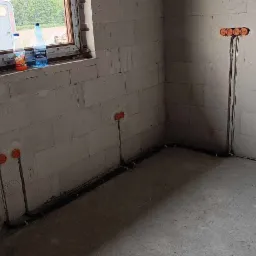 Wnętrze w surowym stanie z widocznymi przewodami elektrycznymi wyprowadzonymi ze ściany, pomarańczowe puszki instalacyjne, przewody ułożone wzdłuż ściany, okno z widokiem na zewnątrz, butelki...