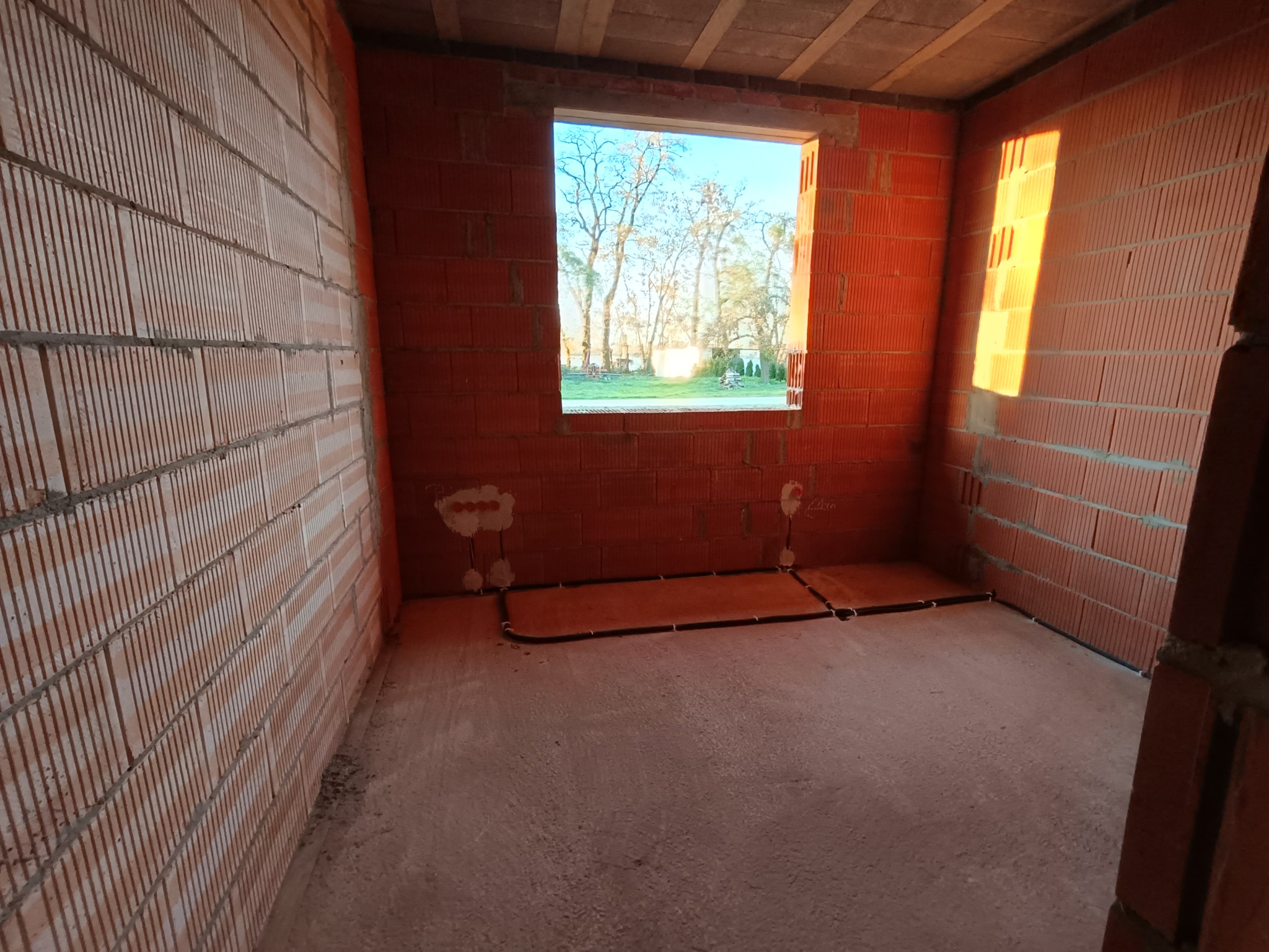 Surowy stan wnętrza budynku w Ostrowite z widocznymi przewodami elektrycznymi ułożonymi przy podłodze, wyprowadzonymi gniazdkami w ścianie z cegły i oknem z widokiem na zieleń.