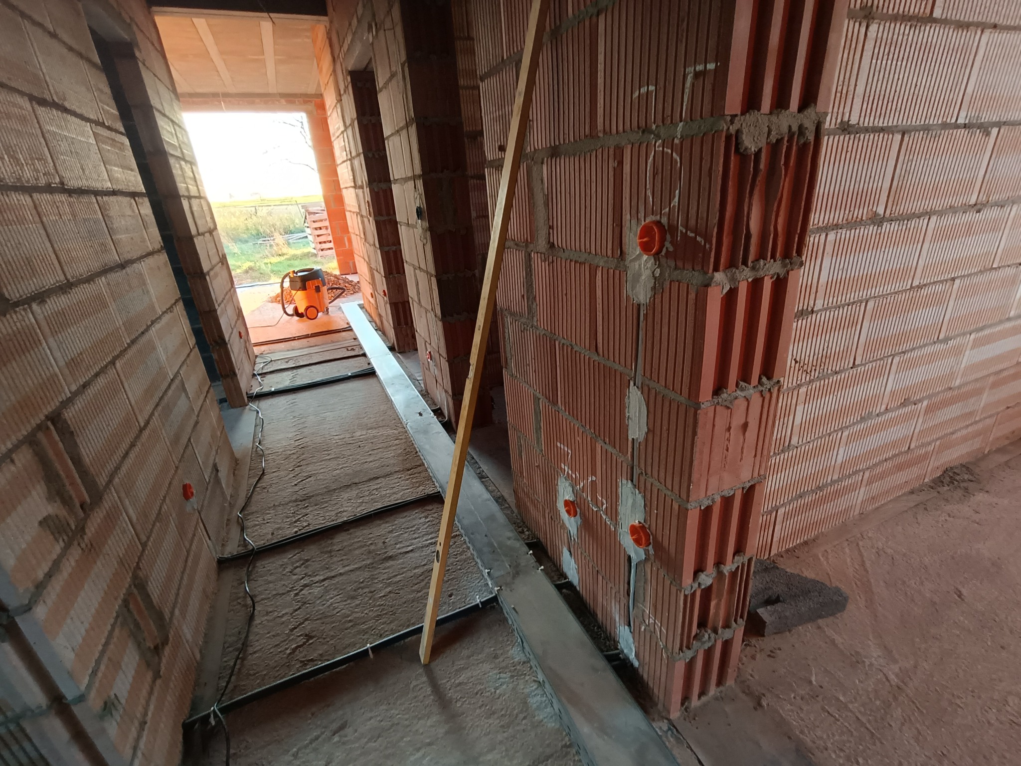 Wnętrze w stanie surowym: ściany z czerwonej cegły dziurawki z widocznymi puszkami instalacyjnymi, poziomica oparta o ścianę, w oddali pomarańczowy odkurzacz przemysłowy.