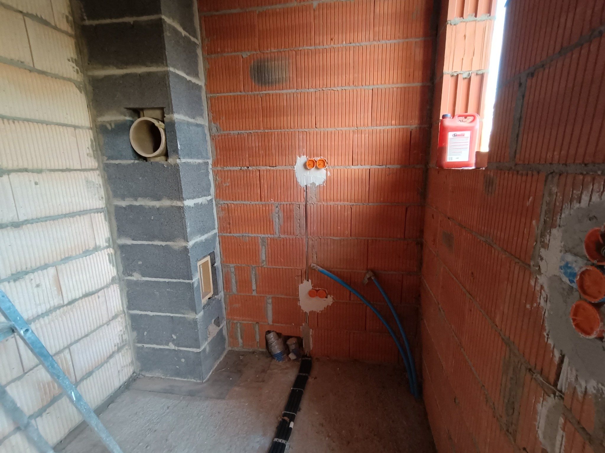 Surowy stan wnętrza w trakcie prac instalacyjnych: widoczne puszki elektryczne zalane gipsem w czerwonej cegle, rury instalacji wodnej, pion kominowy z otworem wentylacyjnym oraz wiązka przewodów...