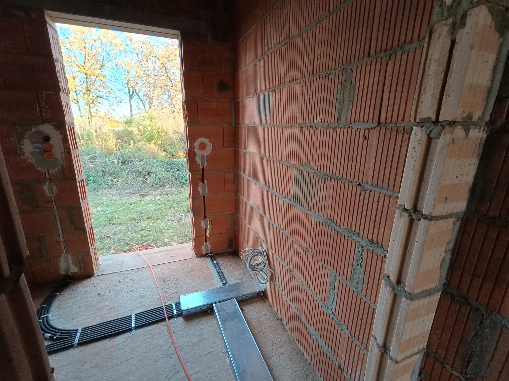 Wnętrze surowego budynku z widoczną instalacją elektryczną: peszle z kablami ułożone wzdłuż ściany z cegły ceramicznej, uchwyty montażowe, puszki instalacyjne w ścianie, widok na zewnątrz przez...