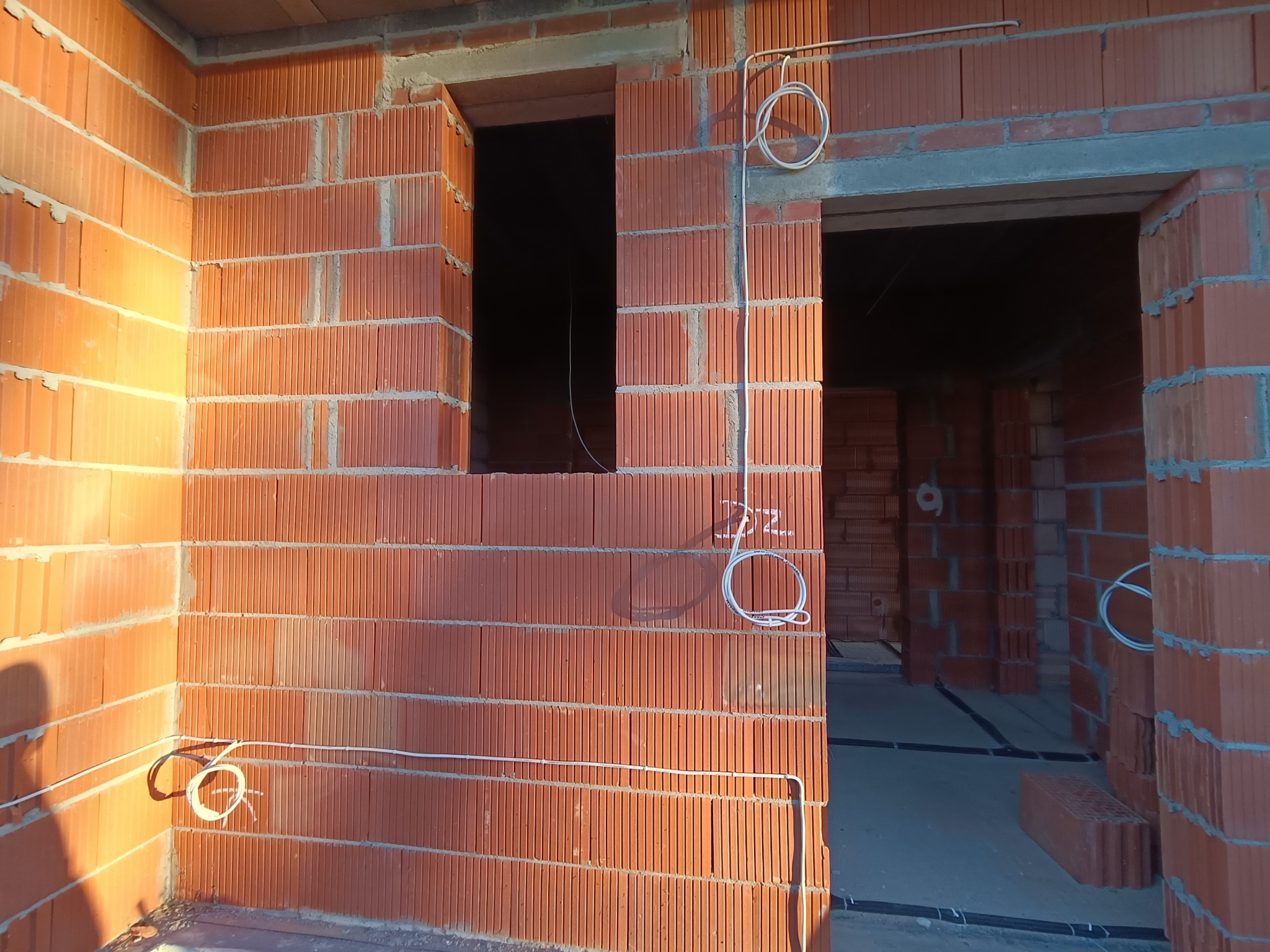 Wnętrze budynku w trakcie budowy z widocznymi przewodami elektrycznymi poprowadzonymi po ścianach z czerwonej cegły dziurawki, kable zwinięte w pętle przy ścianach, otwór okienny i drzwiowy.