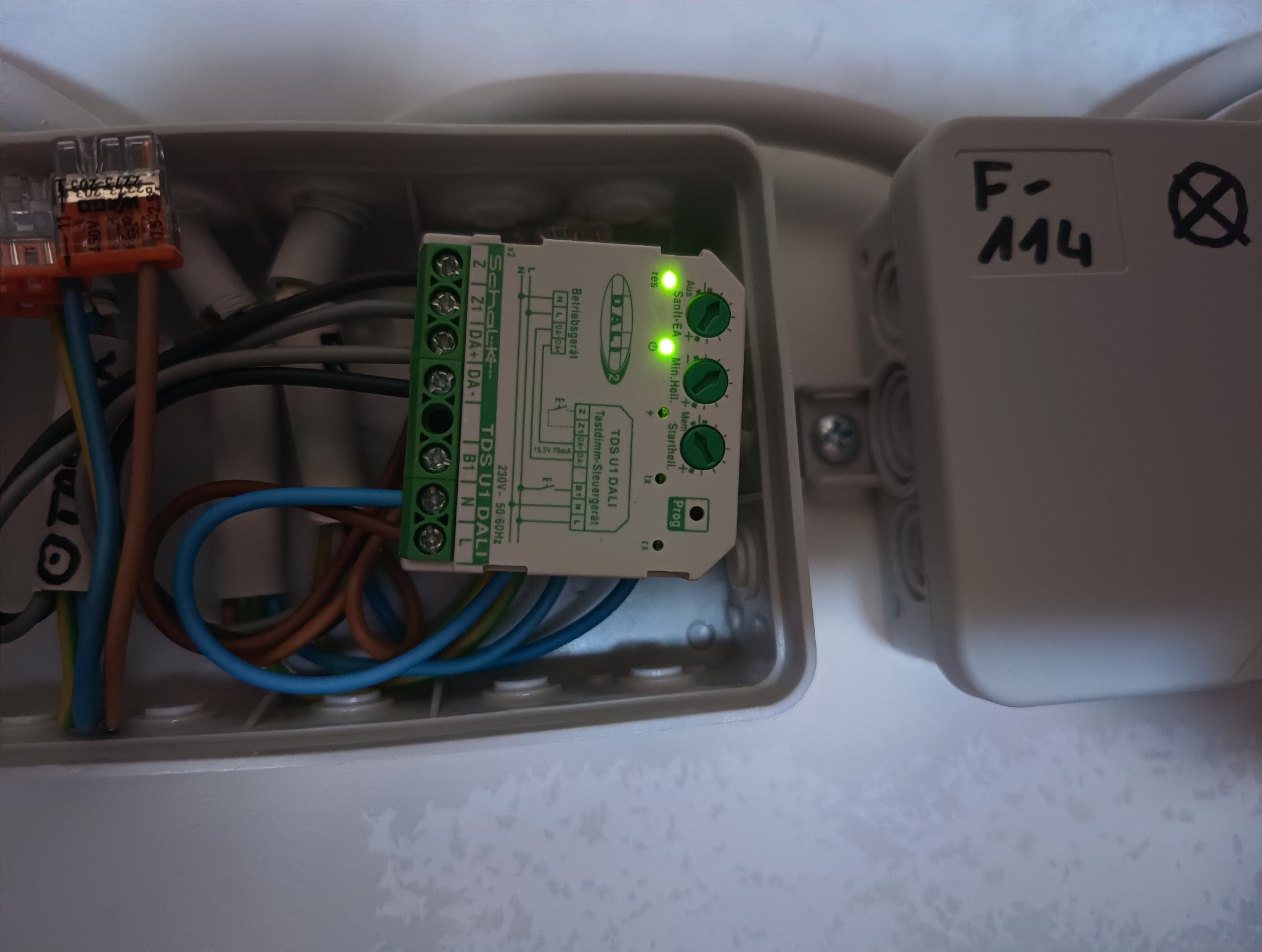 Otwarta puszka elektryczna z kontrolerem DALI Schalk TDS U1, widoczne podłączenie przewodów, złącza WAGO i diody LED sygnalizujące pracę urządzenia. Na sąsiedniej puszce widoczny napis F-114.