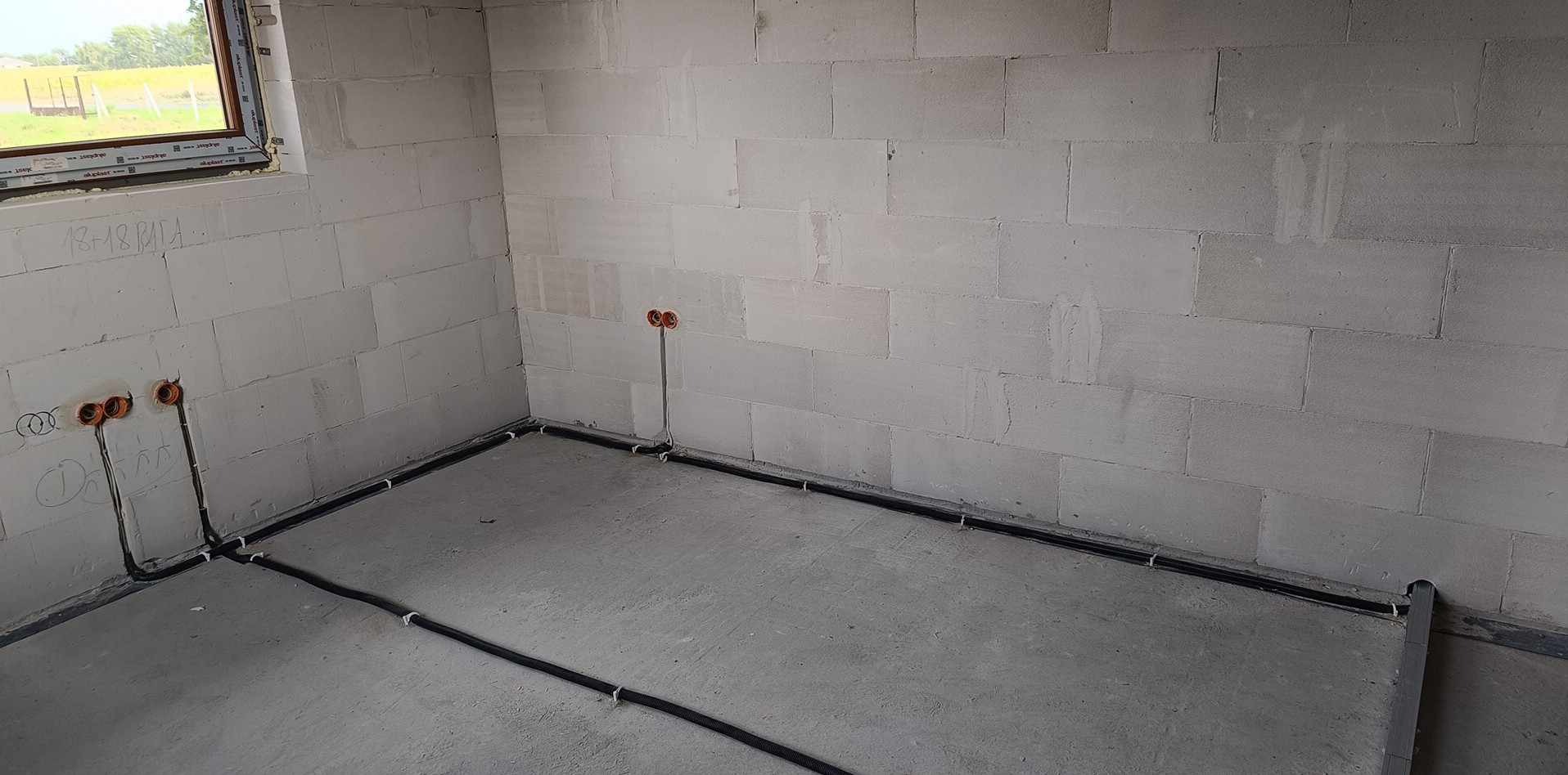 Surowy stan: Czarna instalacja elektryczna w peszlach, przytwierdzona do szarej posadzki i ścian z betonu komórkowego, z widocznymi puszkami instalacyjnymi.