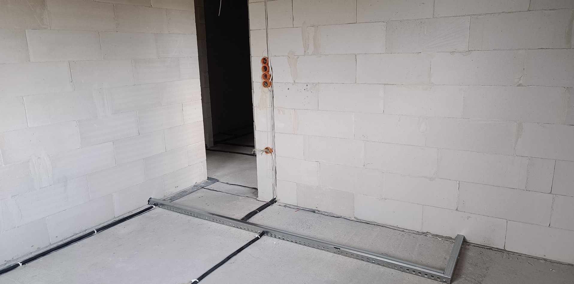 Surowy stan budynku z widocznymi przewodami elektrycznymi wychodzącymi ze ściany, metalowa listwa przypodłogowa, ściany z białych bloczków.