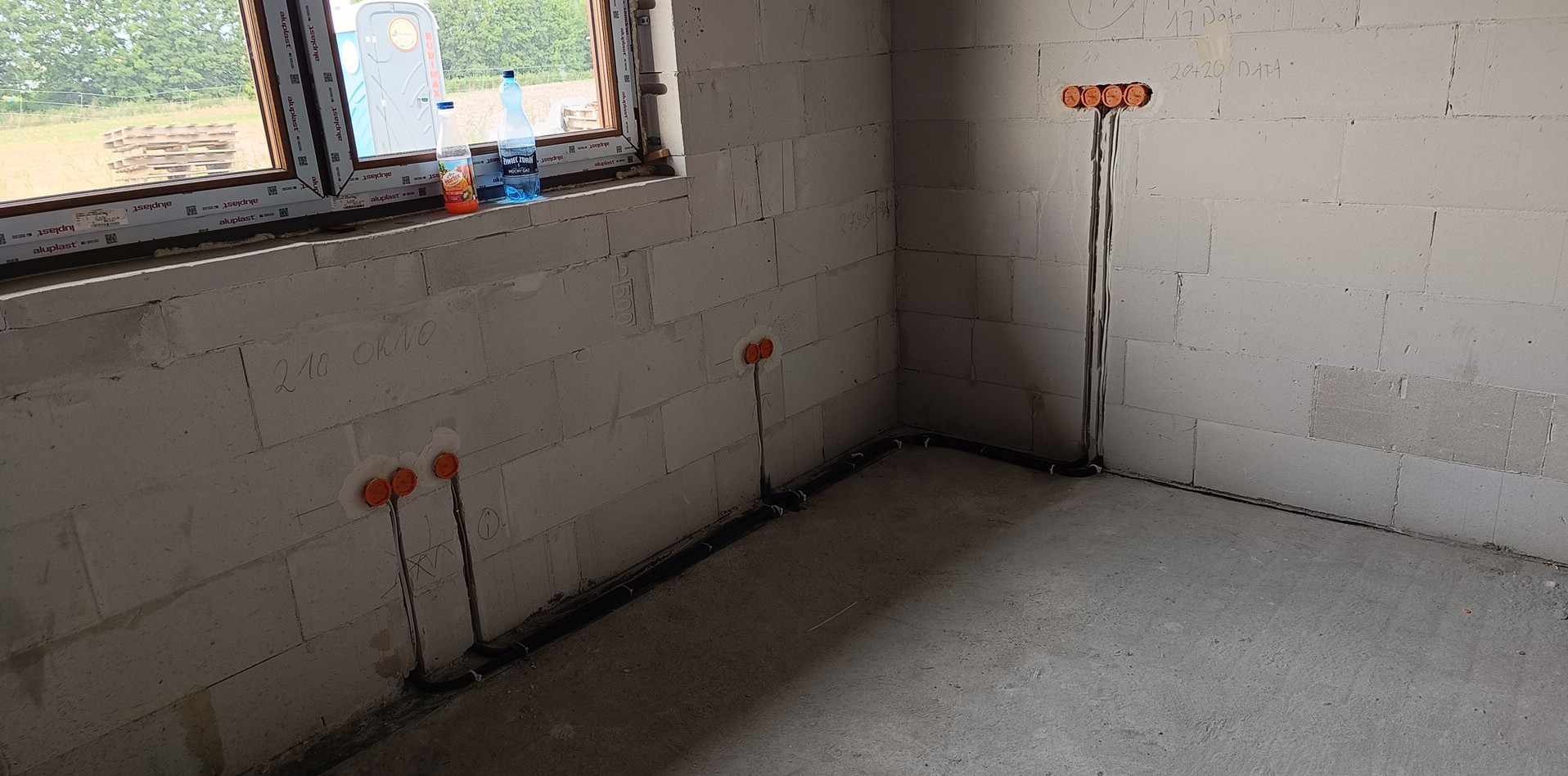 Wnętrze w surowym stanie z widocznymi przewodami elektrycznymi wyprowadzonymi ze ściany, pomarańczowe puszki instalacyjne, przewody ułożone wzdłuż ściany, okno z widokiem na zewnątrz, butelki...