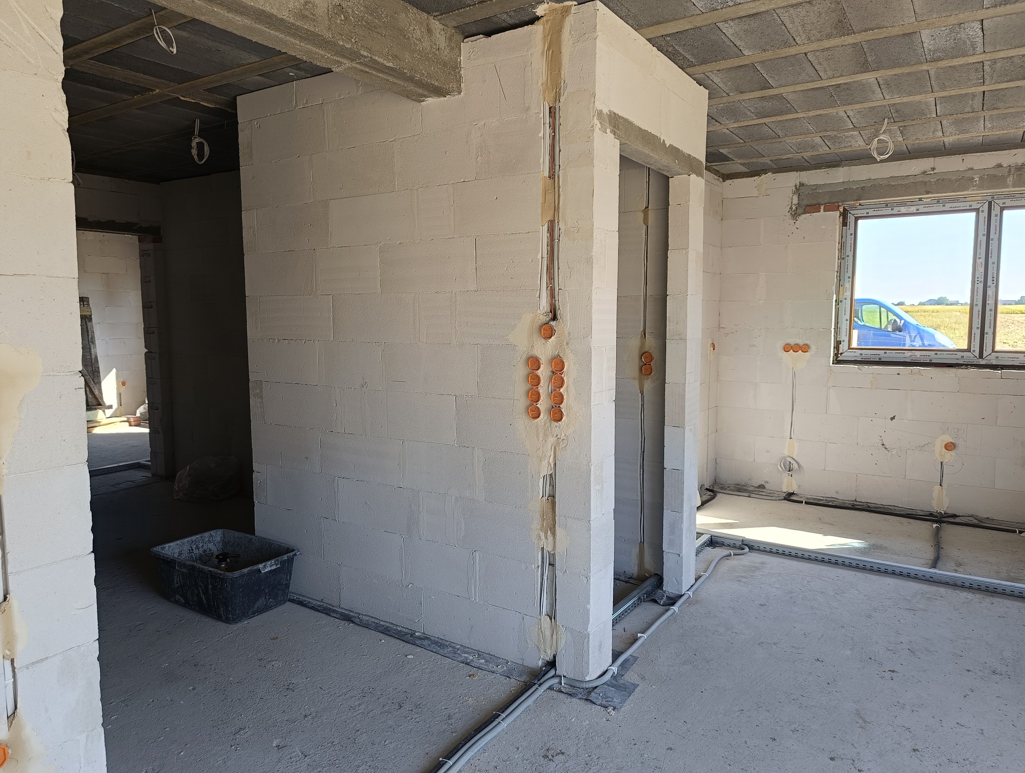 Wnętrze surowego budynku w trakcie prac instalacyjnych: widoczne bruzdy w ścianach z poprowadzonymi przewodami elektrycznymi, puszki instalacyjne, szare rury na podłodze, okno z widokiem na pole.