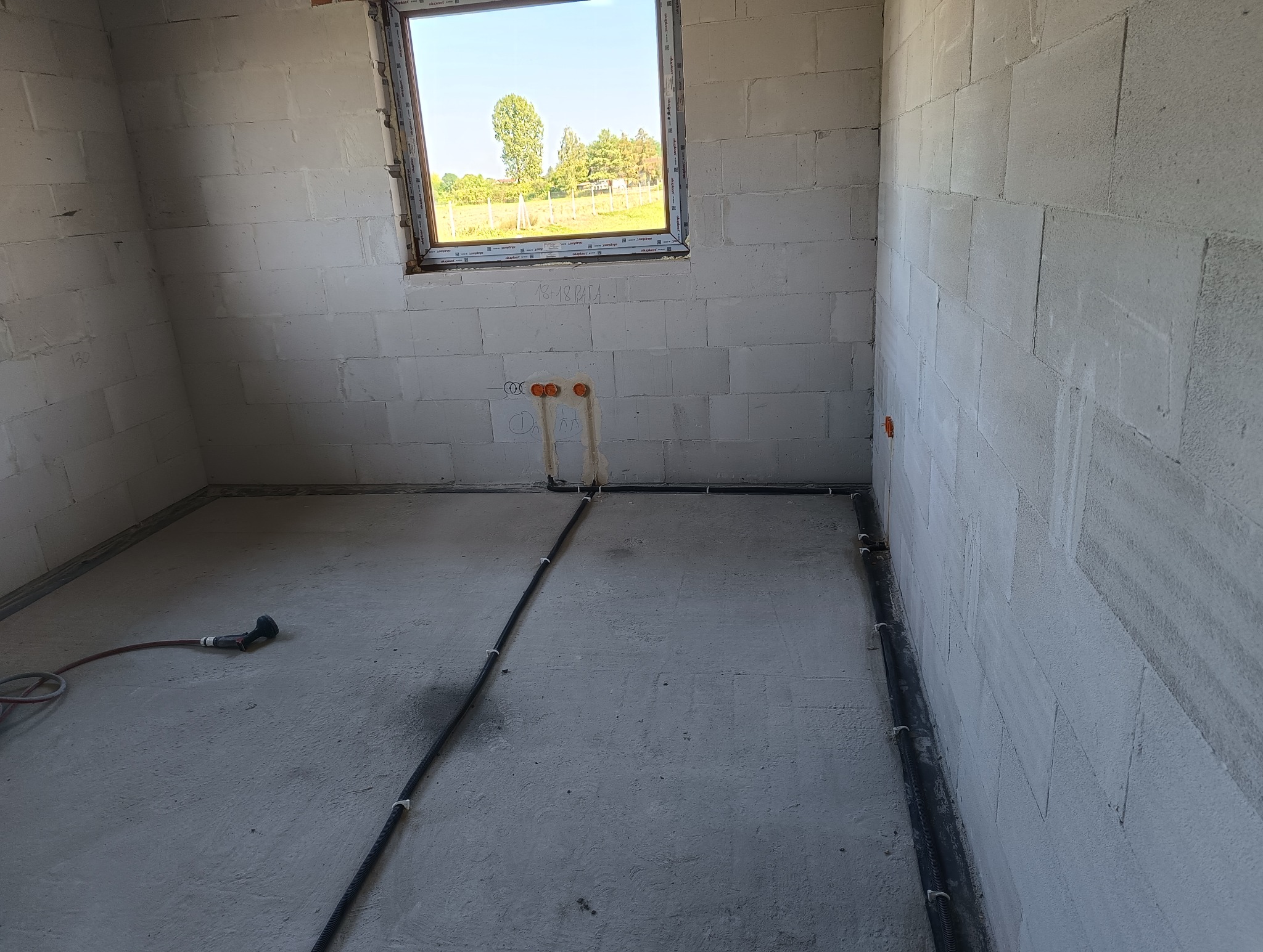Wnętrze surowego budynku w trakcie instalacji elektrycznej: widoczne kable w peszlach przy ścianach i wyprowadzenia przewodów pod gniazdka, tło stanowi okno z widokiem na zewnątrz.