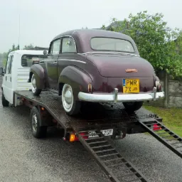 Laweta z bordowym, zabytkowym Oplem Olympia z żółtą tablicą PWR 7X na platformie. Na mokrej jezdni, w tle zieleń.