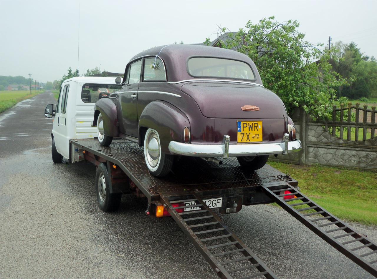 Laweta z bordowym, zabytkowym Oplem Olympia z żółtą tablicą PWR 7X na platformie. Na mokrej jezdni, w tle zieleń.