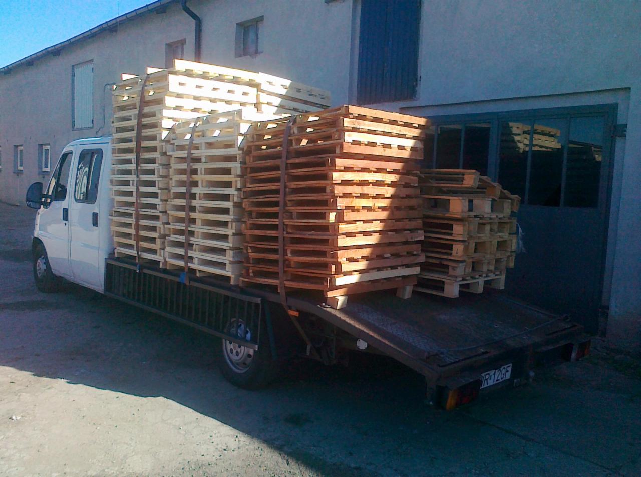 Biały bus z otwartą platformą przewozi jasne i ciemne palety drewniane. Budynek w tle, światło słoneczne.