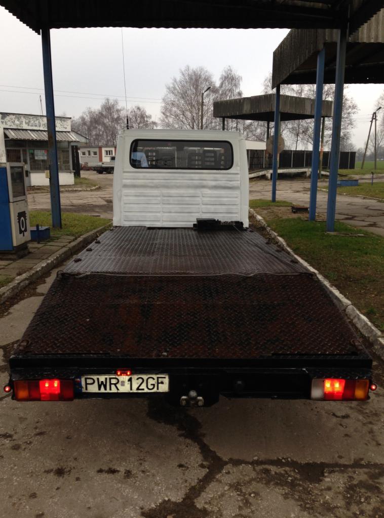 Tył białego busa-lawety z widoczną tablicą rejestracyjną PWR-12GF, stojącego na betonowym placu w pochmurny dzień. Widoczne budynki w tle.