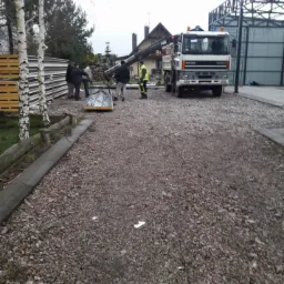 Montaż konstrukcji metalowej przy użyciu dźwigu samochodowego; widoczni pracownicy w odzieży roboczej, teren utwardzony żwirem, w tle budynek mieszkalny i hala.