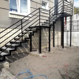 Zewnętrzne schody metalowe z czarnymi poręczami prowadzące do drzwi na piętrze budynku, widoczne przewody elektryczne na ziemi sugerują trwające prace wykończeniowe.